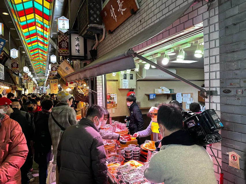 사람들이 다양한 가판대에서 쇼핑하는 분주한 실내 시장. 상인들이 포장된 음식 품목을 포함한 다양한 상품을 판매하고 있다. 많은 쇼핑객과 상인들이 얼굴 마스크를 착용하고 있다. 천장은 다채로운 스테인드 글라스 패널로 장식되어 있어 장면에 생동감 있는 요소를 더하고 있다.