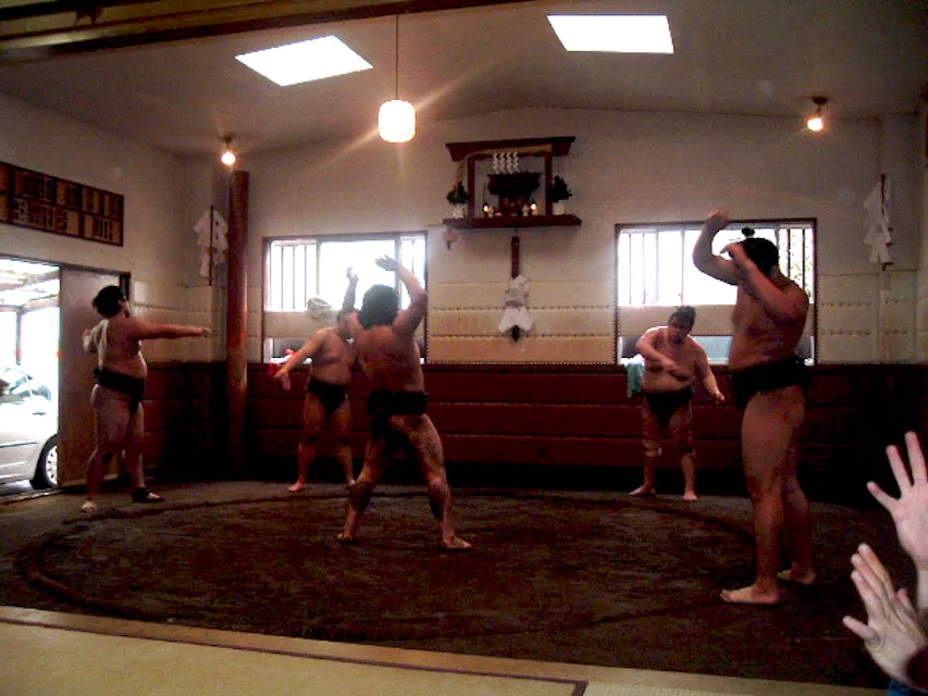 Inside a Sumo Stable 스모 선수들이 흙 링이 있는 훈련 마구간에서 훈련하고 있습니다. 전통적인 마와시 벨트를 착용한 네 명의 선수들이 몸 풀기 운동을 하고 있으며, 다른 한 사람은 사이드라인에서 지켜보고 있습니다. 기도 로프와 의식 장식이 방을 장식하고 있습니다.