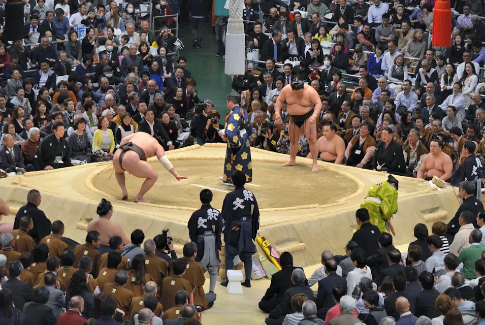 Sumo Wrestling 모래 링에서 큰 관중이 둘러싼 가운데 스모 레슬링 경기이 열리고 있다. 두 명의 스모 레슬러가 경기 시작 준비를 하며 서로 마주 보고 서 있고, 전통 복장을 한 심판이 그들 사이에 서 있다. 많은 관중들이 집중해서 보고 있다.