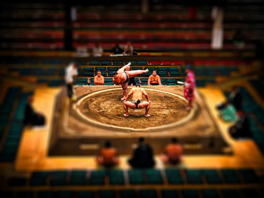 Sumo Wrestling 두 명의 스모 레슬러가 링에서 경쟁하며 한 선수가 다른 선수를 들어 올리고 있습니다. 아레나는 비어 있는 좌석 행으로 둘러싸여 있고, 링 구역 내에서 몇 명의 사람들이 경기를 관람하며 집중되고 강렬한 분위기를 더하고 있습니다.