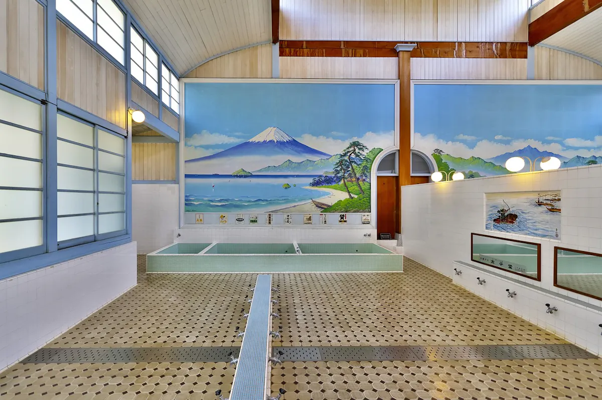 A traditional Japanese bathhouse interior with tile flooring, a mural of Mount Fuji on the wall, wide windows, and light fixtures. There are several washing stations with mirrors and faucets, along with communal soaking pools.