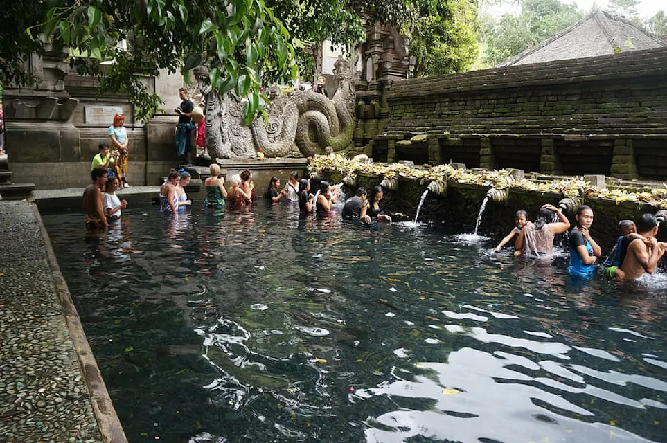 Japanese Public Bath A group of people stand in a temple's stone pool, beneath multiple small water spouts. The area is surrounded by greenery and stone sculptures, including a large serpent statue. Visitors are seen bathing and gathering in the serene, sacred setting.