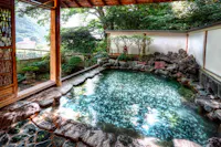 An inviting outdoor onsen (hot spring bath) with clear, steaming water surrounded by natural rocks. A wooden pergola and lush greenery enhance the tranquil atmosphere, with a white wall and trees providing privacy and a sense of seclusion.