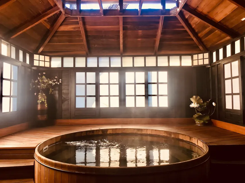 Rejuvenating Onsen A serene indoor hot spring bathhouse featuring a wooden circular tub filled with steaming water. The room is adorned with dark wooden panels and paper-covered windows, allowing soft natural light to filter through. A potted plant and a flower vase add a touch of elegance.