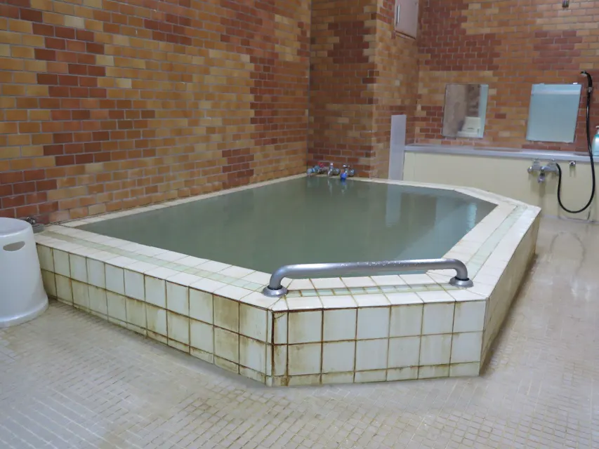 Japanese Public Bath A rectangular indoor bathing area with tiled steps and handrails, partially filled with water. The surrounding walls are covered in tan and brown bricks. The area includes a shower hose, faucet, and mirror. The floor has square tiles with a drain and small stool nearby.