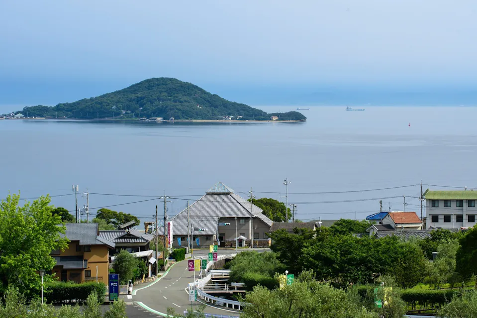 静かな沿岸の町の眺め、背景に丘の多い島があり、穏やかな青い海と晴れた空。前景には家々、緑の木々、そして曲がりくねった道路が描かれています。この光景は平和で絵のように美しく、魅力的な海辺の雰囲気を反映しています。