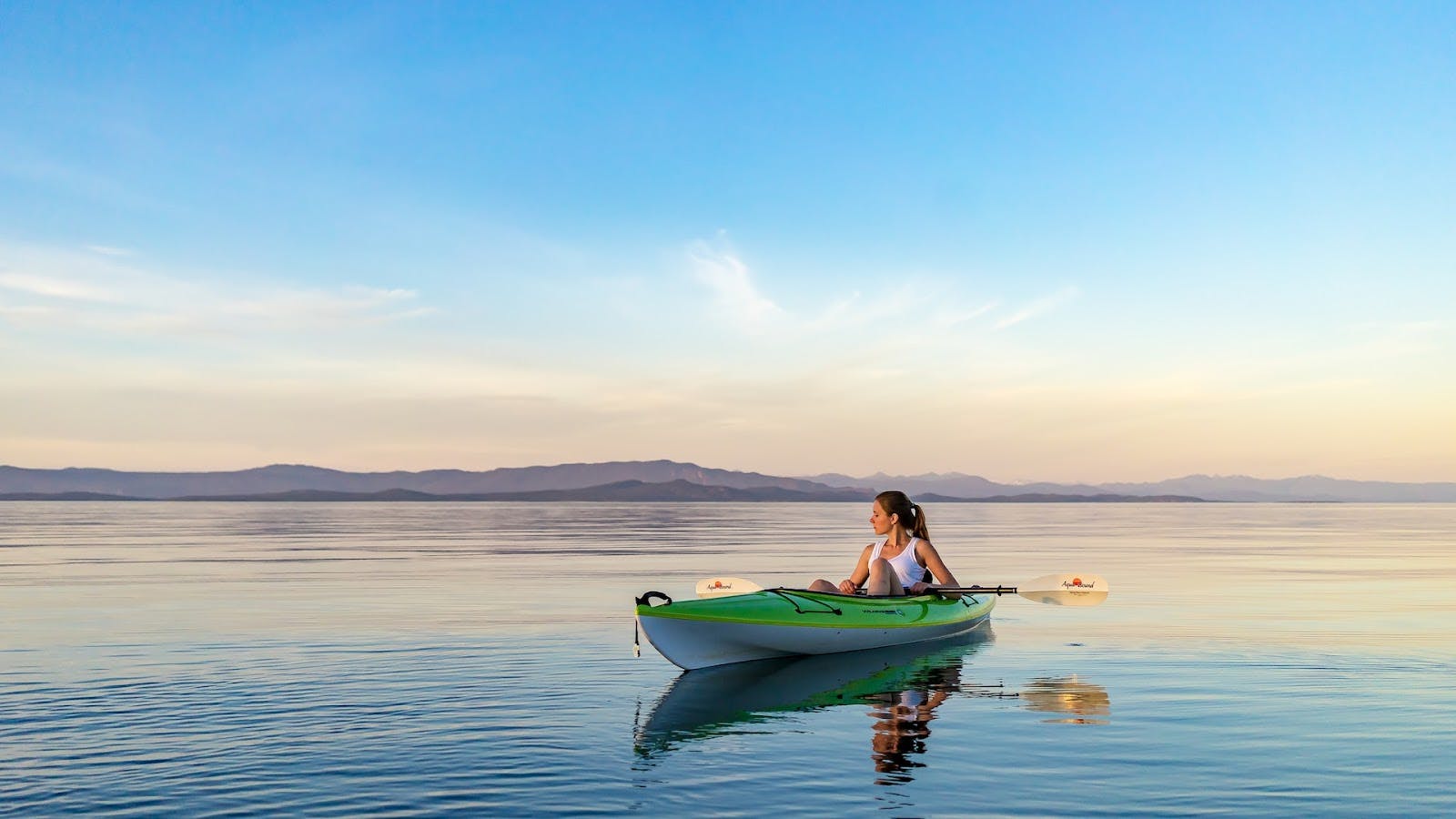 A person in a green kayak paddling on a calm lake during sunset. The sky is clear with a few wispy clouds, and mountains can be seen in the distance. The water reflects the pastel colors of the sky, creating a serene and peaceful scene.