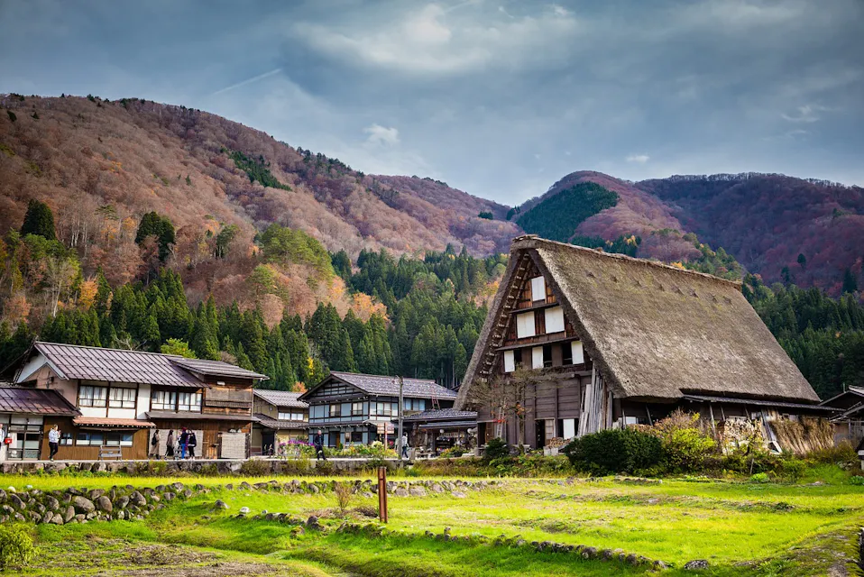 A serene village scene with traditional thatched-roof houses set against a backdrop of forested mountains. The sky is partly cloudy, and vibrant green grass contrasts with the autumn foliage on the hills. A few people are visible, leisurely walking through the village.