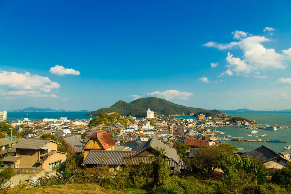 A scenic view of a coastal Japanese town with traditional houses, nestled between lush green hills and blue ocean waters. The clear blue sky is dotted with a few white clouds, and boats are moored at the harbor. The landscape is vibrant and peaceful.