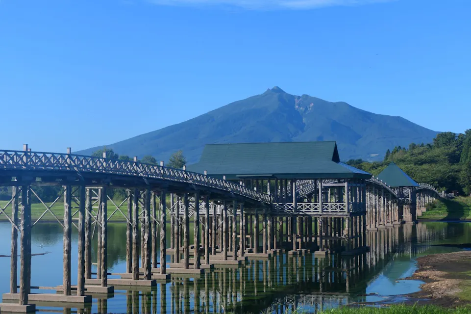 A wooden bridge with multiple covered sections stretches over a calm body of water with tall pillars reflected in the water below. Behind the bridge, a lush green landscape extends towards a tall, prominent mountain peak under a clear blue sky.