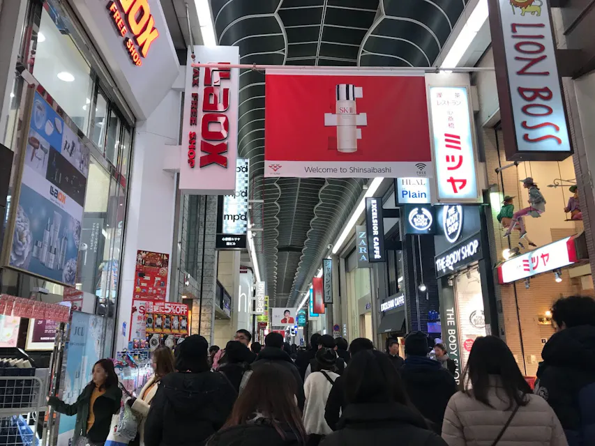 A bustling indoor shopping street is filled with people. Various brightly lit storefront signs in different languages, including Japanese and English, line both sides. A large overhead banner reads "Welcome to Shinsaibashi." The atmosphere is lively and energetic.