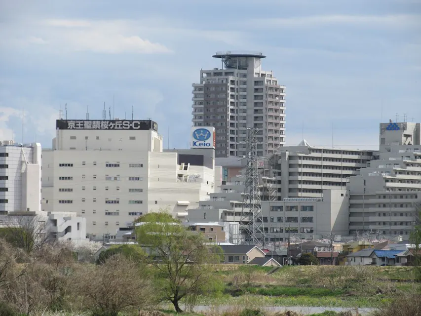 Urban view of a city featuring modern high-rise buildings, including prominent structures with the "Keio" and "Ntt" logos. In the foreground, there is a mix of smaller buildings and greenery, including some trees and a grassy area. A partly cloudy sky is above.