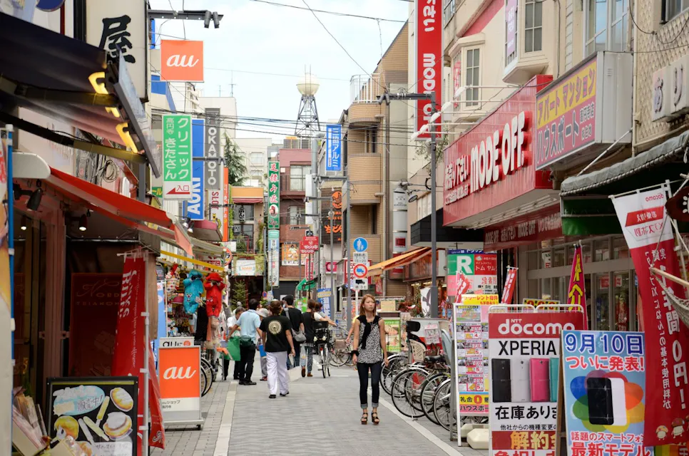 A bustling street scene in a Japanese shopping district with various colorful shop signs and banners. People walk along the street, which is lined with bicycles and shop displays. A tower is visible in the distance.