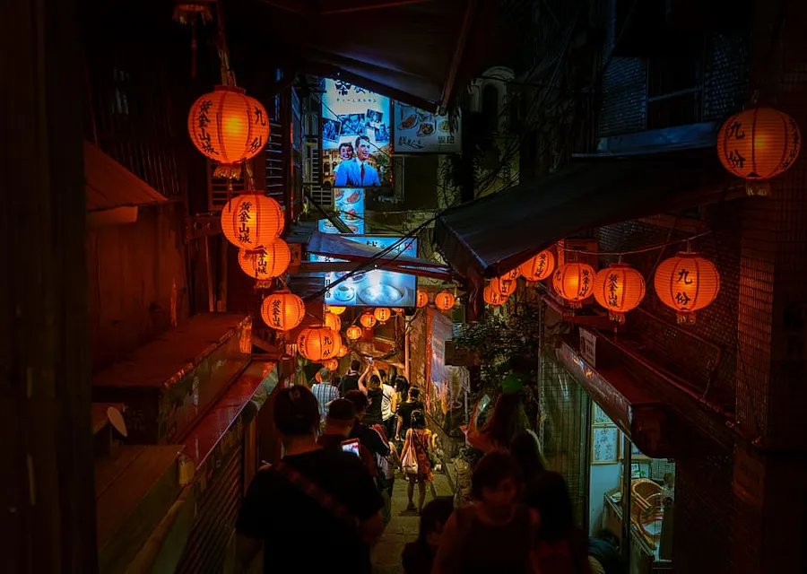 A narrow alley lit by numerous red lanterns with black calligraphy, winding down past multiple traditional buildings. People are descending the steps, while a brightly lit shop at the end of the alley attracts attention.