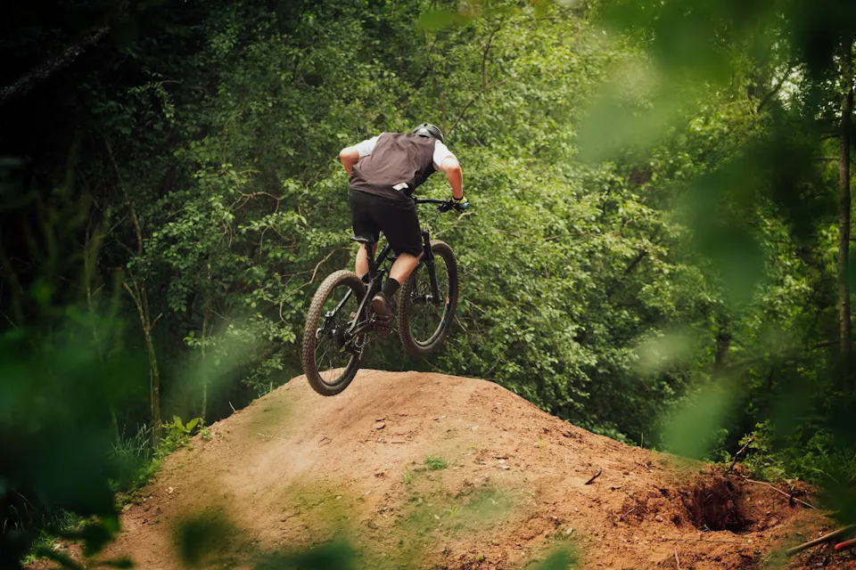 一个穿着头盔、运动衫和短裤的人正在森林区域的土堆上骑山地自行车。这个骑自行车的人在空中，展示出一个跳跃，背景是郁郁葱葱的树木和植被。