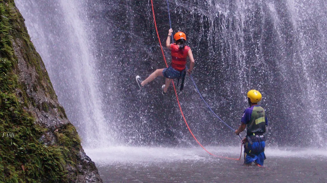 Two people are engaged in waterfall rappelling. One person is hanging mid-air, wearing a red helmet, life vest, and harness, while the other person steadies them from below, wearing a yellow helmet and blue vest. Water cascades behind them, and a moss-covered rock is on the left.