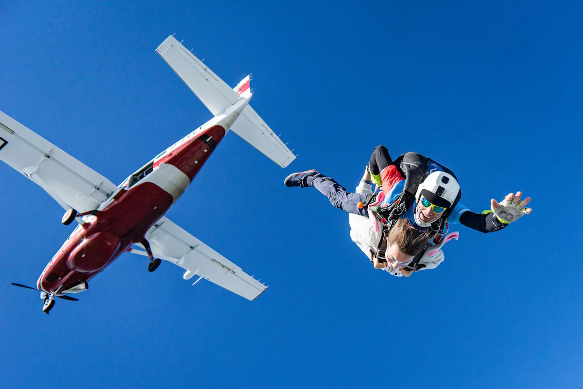 A skydiving instructor and a tandem skydiver freefall beneath a small airplane. The instructor, wearing a white helmet and sunglasses, holds the tandem skydiver, who is in a white jumpsuit. The background features a clear blue sky.