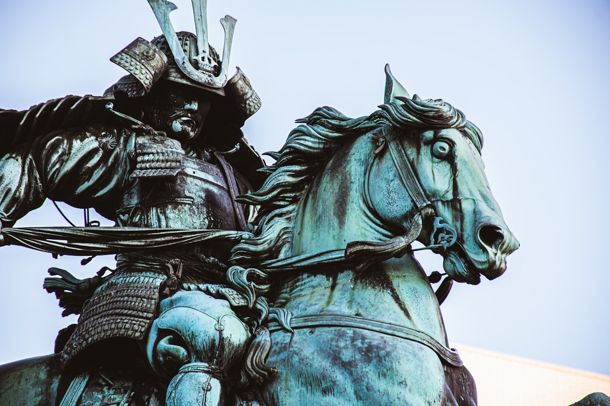 Samurai Bronze statue of a Japanese samurai warrior in traditional armor, riding a rearing horse. The statue is detailed, showcasing the warrior's helmet with an elaborate crest, and the horse's flowing mane. The background features a clear, light blue sky.