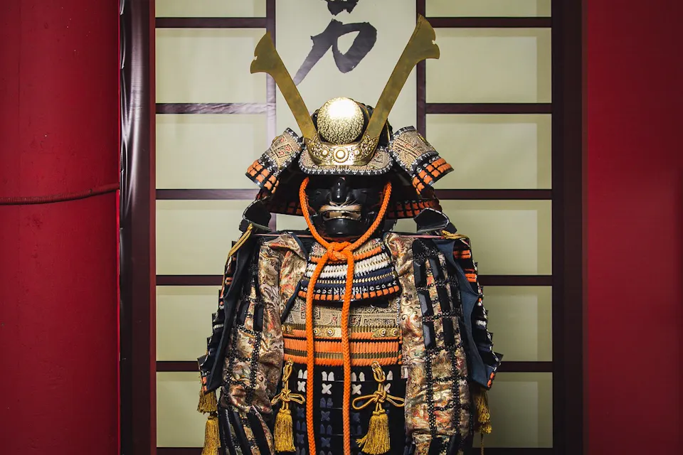 Samurai Armor 一套完整的传统日本武士盔甲,带有大型金色角的头盔,精致的黑色和金色细节,以及橙色绳索装饰,展示在带有日本书法的背景前。盔甲具有华丽且历史悠久的设计。