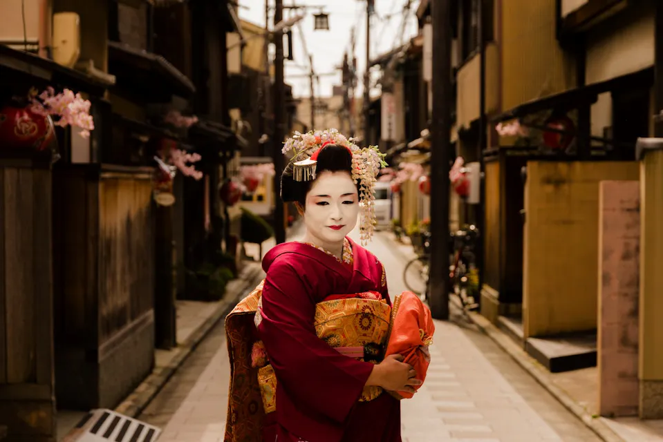 A person in traditional Japanese attire stands in a narrow, old-fashioned street. They wear a richly decorated red kimono and an ornate hairstyle adorned with flowers, embodying the elegance of traditional Japanese culture. The street is lined with weathered buildings.