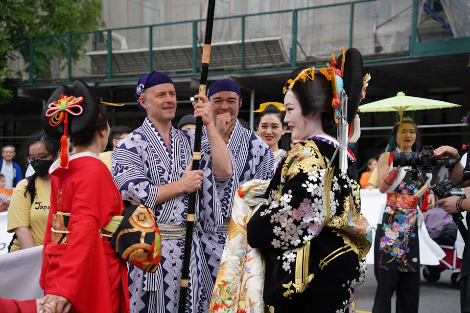A group of people participates in a Japanese cultural festival. Two men in striped traditional garments and headbands hold a tall pole. Four women wearing elaborate kimonos and hairpieces stand near them, one holding a camera. Spectators and a building are in the background.