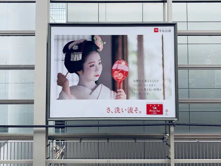 Geisha Poster 一块广告牌位于建筑前面,展示了一位身着传统服饰的艺妓手持红色棒棒糖的形象。广告牌上附有日文文本。背景展示了一座现代的钢铁玻璃结构。