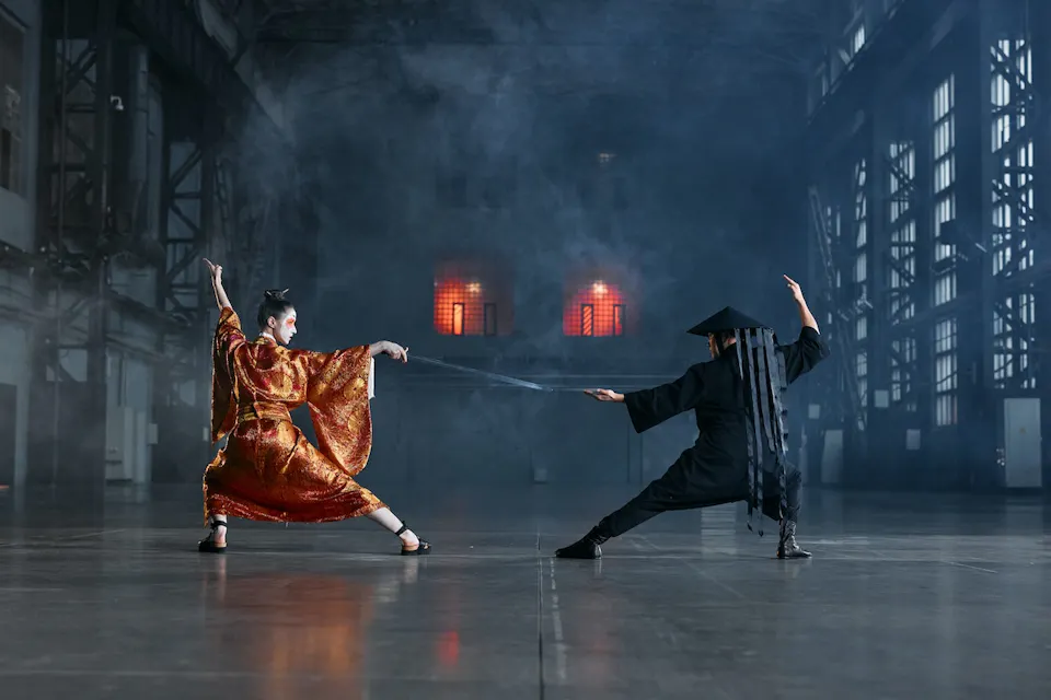 Geisha 两个身穿传统服装的人正在宽敞、昏暗的工业空间内表演戏剧性的战斗场景。其中一人穿着橙色长袍,手持武士刀,另一人则穿着黑色长袍,伸出手臂防御,形成了动态的视觉对比。