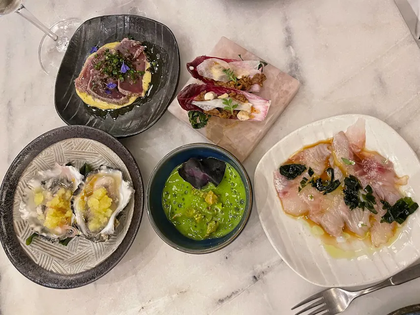Ryugin 一盘精美摆放的美食选择在大理石桌子上,包括黑盘上的煎金枪鱼、生蚝、碗中的绿色汤、牛肉薄片,以及一种用红菊苣叶包裹奶油馅料的菜肴。