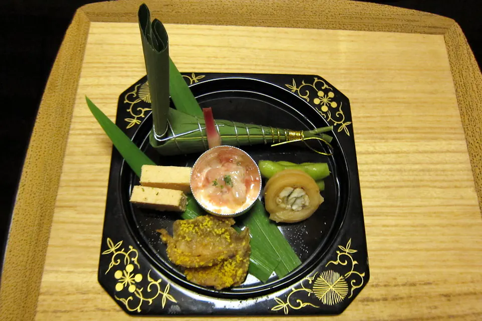 Kashiwaya 一盘精美摆放的日本菜肴,盛放在一个黑色漆面盘子上,盘子上有精致的金色图案。盘子里有各种食物,包括包裹的食材、一个酱汁碟、豆腐块、一个圆柱形食物和装饰,所有食材与绿叶优雅地呈现。