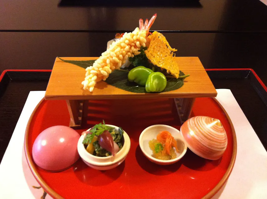 Nakamura 精心安排的日本餐:在高架木托盘上盛放的天妇罗虾和玉米,搭配葱花,摆放在一个红色托盘上,配有侧菜,包括一个粉色的蛋,绿色的蔬菜配料,一个贝壳,以及一个盛有切片肉和蔬菜的小碗。
