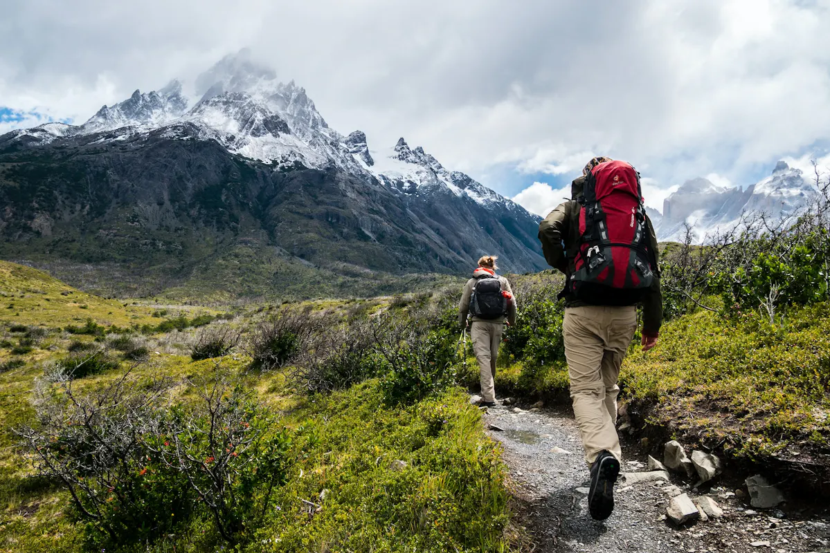 Two hikers walk along a narrow dirt trail surrounded by lush green bushes, with snow-capped mountains towering in the background under a cloudy sky. Both are wearing backpacks, with the lead hiker in a red backpack and the second in a gray one.