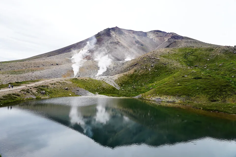 一幅宁静的山脉景观，湖水平静，映照出山峰和周围景色。蒸汽口在山顶附近释放出白色烟雾。前景为郁郁葱葱的绿草，向岩石地形过渡，通往山脉。