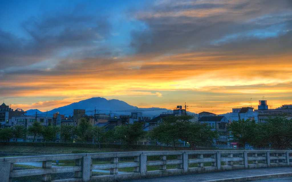 一轮充满活力的日落在远处的山脉后面，给小镇披上了一层金色的色调。天空中有蓝色、黄色和橙色的条纹，而小镇由低层建筑和树木组成。前景中可见一座混凝土桥。