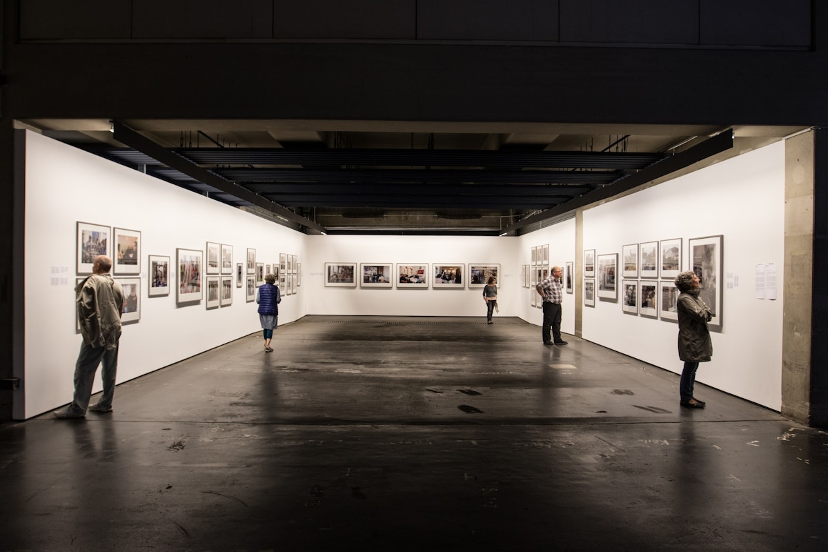 Art Gallery A spacious art gallery with white walls showcasing framed artworks. Several people are observing the pieces, maintaining a distance. The room is well-lit, emphasizing the displayed art. The floor is dark, contrasting with the bright gallery space.