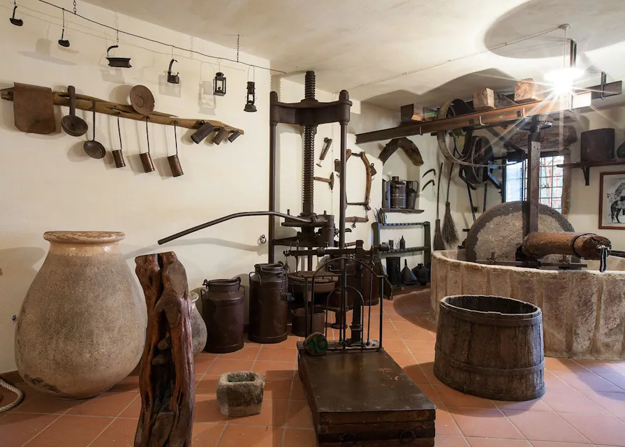 전통 와인과 올리브 오일 생산에 사용되는 역사적 도구와 유물이 가득한 방. 이 공간에는 큰 점토 항아리, 나무 통, 금속 조리기구, 압착 장비 및 분쇄기 돌이 포함되어 있으며, 모든 것이 타일 바닥과 벽에 깔끔하게 배열되어 있습니다.
