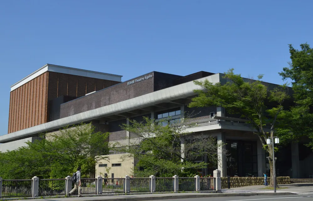 벽돌과 콘크리트 외관을 가진 현대식 건물로, 큰 창문이 있고 나무로 둘러싸여 있습니다. 한 사람이 앞쪽 인도에서 걷고 있으며, 맑고 푸른 하늘이 위에 있습니다. 이 건물은 극장이나 유사한 공공 시설로 보입니다.