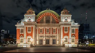 A grand, illuminated red and white brick building with symmetrical architecture stands against a night sky. It features archways, columns, decorative elements, and two towers on either side, with city buildings in the background.
