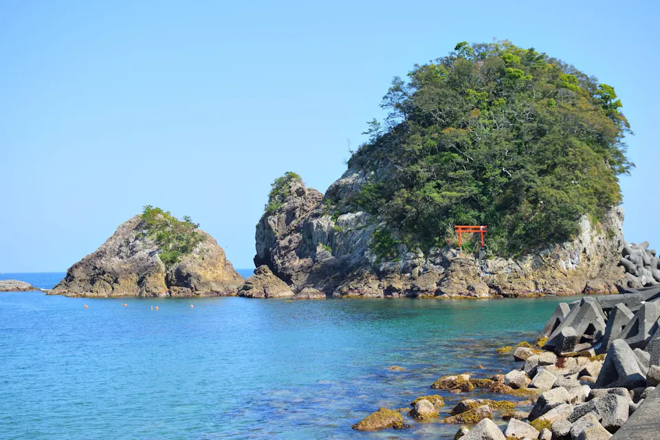 A serene coastal scene featuring an azure ocean with two rocky islets. One islet has a traditional red torii gate nestled among green trees. The foreground includes hexagonal concrete blocks along the shore. The sky is clear and blue, enhancing the tranquil atmosphere.