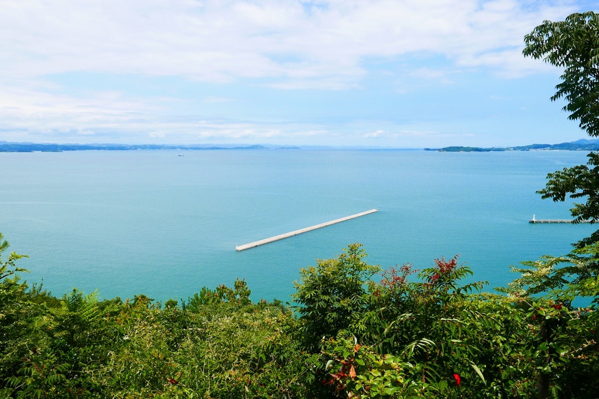 Sea in Japan A panoramic view of a calm blue sea with a long, narrow breakwater extending into the water. The foreground features lush, green vegetation, while a clear sky with scattered clouds stretches over the horizon in the background. Distant landmasses are visible.