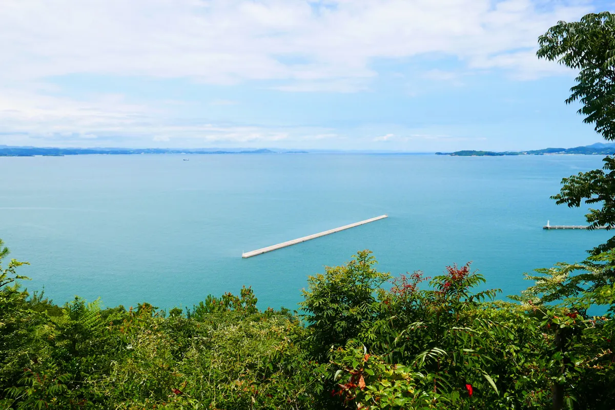 Sea in Japan A panoramic view of a calm blue sea with a long, narrow breakwater extending into the water. The foreground features lush, green vegetation, while a clear sky with scattered clouds stretches over the horizon in the background. Distant landmasses are visible.