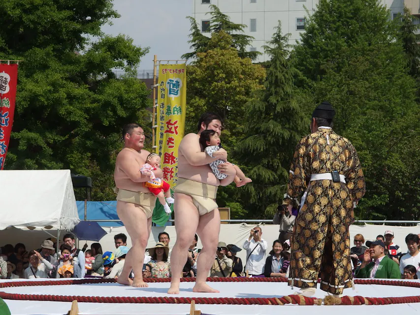 相扑选手每人抱着一个婴儿，站在相扑擂台上参加活动，而一位身着传统服装的官员正在面对他们。背景是树木和五彩斑斓的横幅，观众在后面观看。