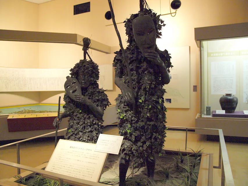 一个博物馆展览，展示了两个身穿黑暗、叶子状服装并佩戴面具的人物，代表一种传统文化实践。前景有一个详细的信息牌，而背景中可见一个模型和陈列着文物的橱窗。