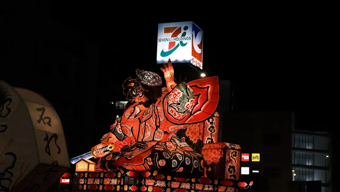 一个生动的、照亮的花车展示了一位手持剑的凶猛战士，夜间展出。这个花车设计精美，颜色鲜艳。在背景中，可以看到一个明亮的“七&I控股”标志。还可以看到其他小型灯光和标志。