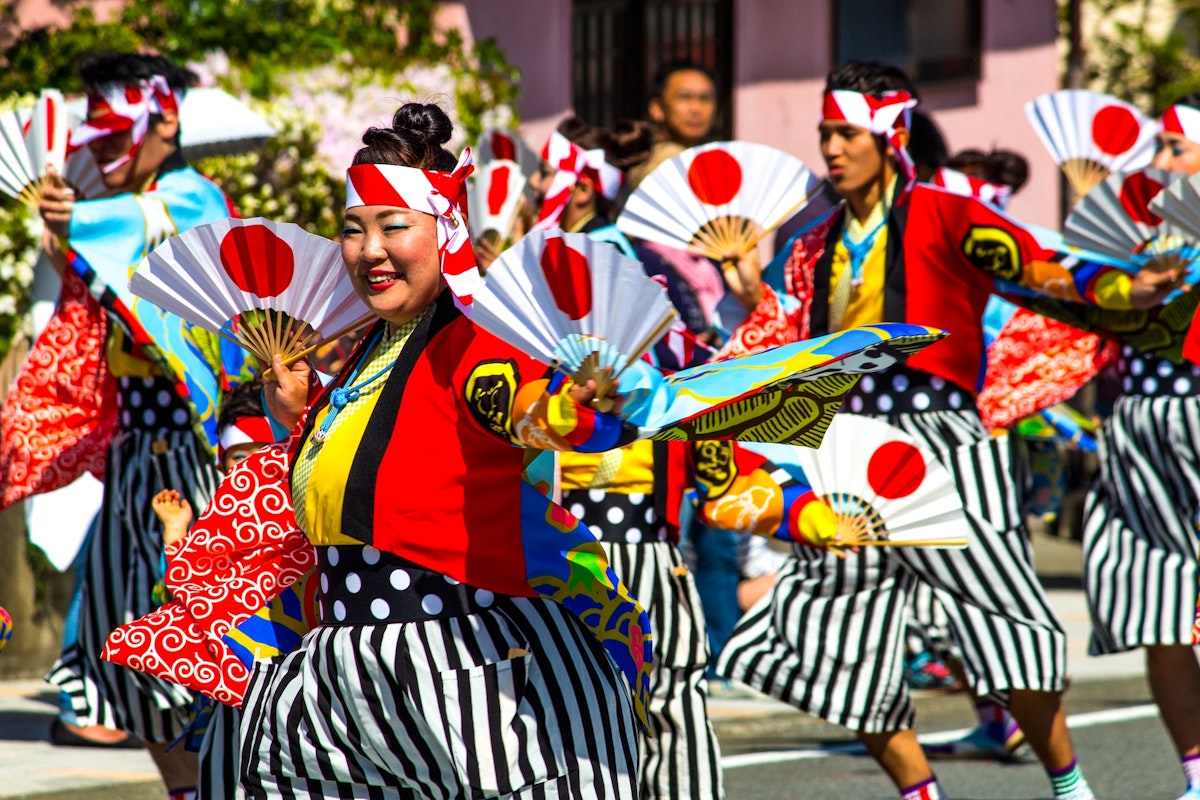 Japanese Festival A vibrant group of people wearing colorful traditional Japanese clothing and headbands perform in a parade. They are energetically dancing and holding decorative fans featuring the Japanese flag. The scene is festive, with smiles and synchronized movements.