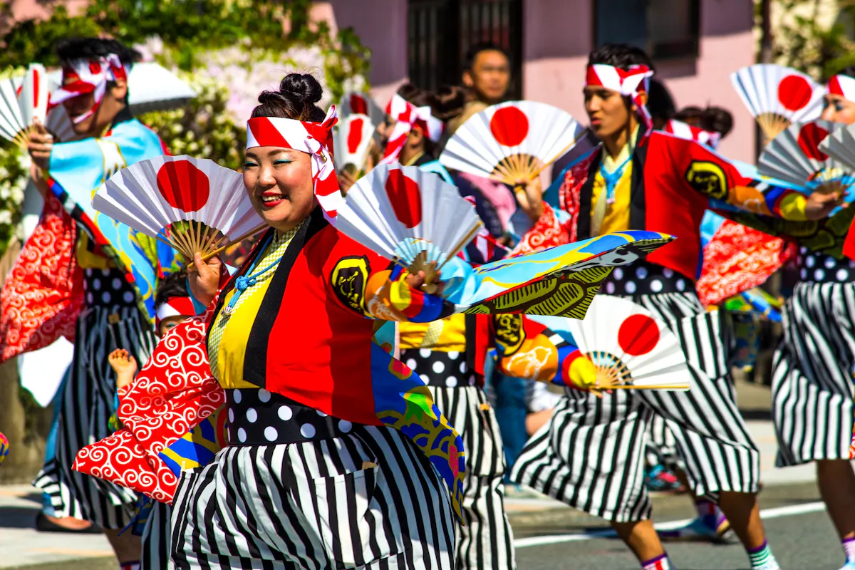 A vibrant group of people wearing colorful traditional Japanese clothing and headbands perform in a parade. They are energetically dancing and holding decorative fans featuring the Japanese flag. The scene is festive, with smiles and synchronized movements.