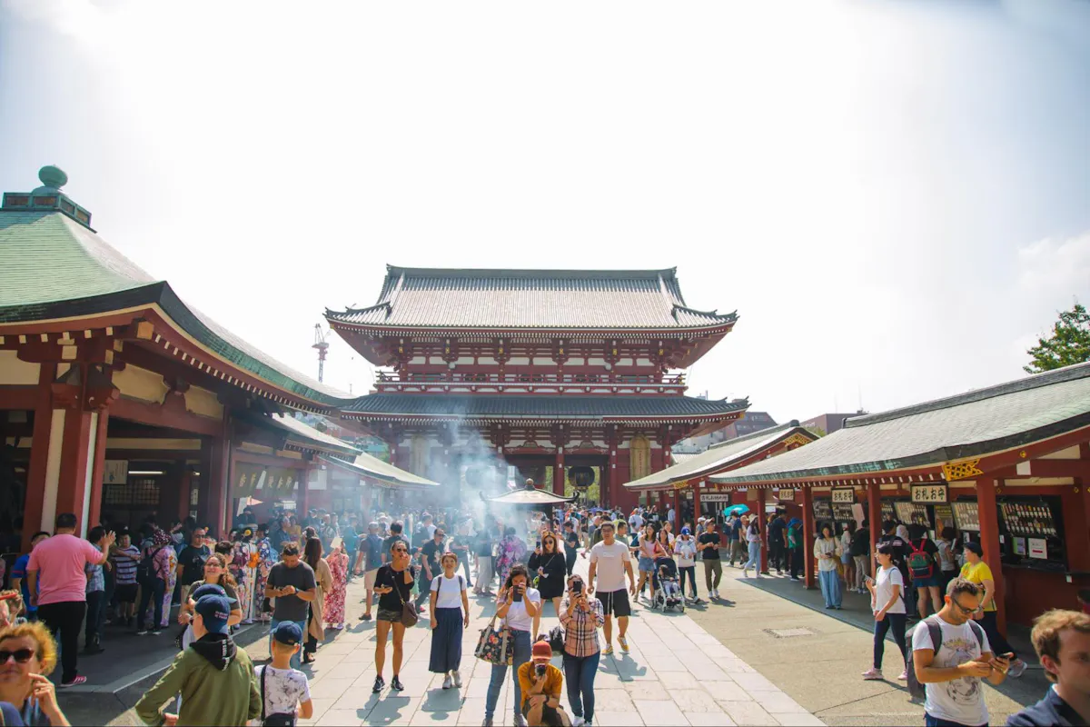 Asakusa A bustling scene at Senso-ji Temple in Tokyo, Japan, with a large crowd of visitors walking through the temple grounds. The main hall, with its traditional architecture, stands prominently in the background, partially obscured by smoke from incense burning.