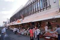 A bustling outdoor market with numerous stalls selling various goods, including seafood and produce. Shoppers browse and interact with vendors under a canopy extending from the building. Signage with Asian characters and bright colors is visible above the stalls.