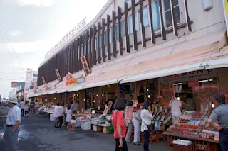 A bustling outdoor market with numerous stalls selling various goods, including seafood and produce. Shoppers browse and interact with vendors under a canopy extending from the building. Signage with Asian characters and bright colors is visible above the stalls.