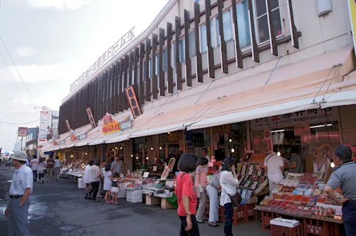 A bustling outdoor market with numerous stalls selling various goods, including seafood and produce. Shoppers browse and interact with vendors under a canopy extending from the building. Signage with Asian characters and bright colors is visible above the stalls.