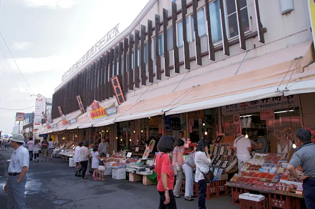 A bustling outdoor market with numerous stalls selling various goods, including seafood and produce. Shoppers browse and interact with vendors under a canopy extending from the building. Signage with Asian characters and bright colors is visible above the stalls.